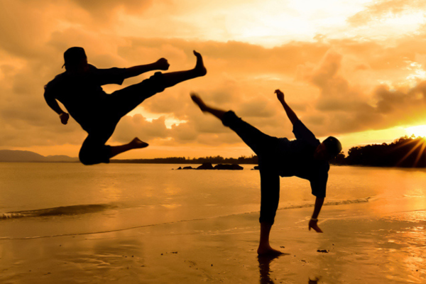 Twee martial artists oefenen trappen op een strand tijdens zonsondergang, met dramatische silhouetten tegen een oranje lucht en reflectie op het natte zand.