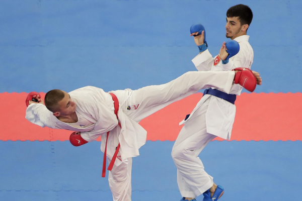 Twee karateka’s in witte gi’s tijdens een wedstrijd; de één voert een hoge trap uit terwijl de ander in verdedigingshouding staat.