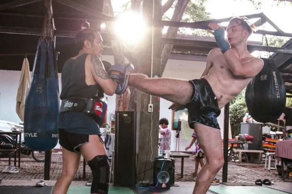 Alt-tekst: Twee mannen trainen Muay Thai in een openlucht gym; de ene geeft een hoge trap terwijl de ander de trap opvangt met pads, met zonlicht dat door het dak schijnt.