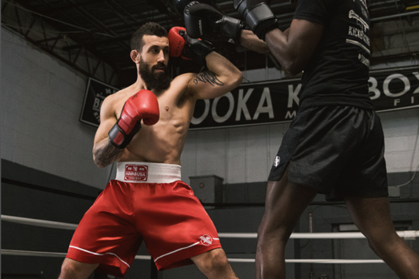 Twee kickboksers in de ring oefenen een combinatietraining, waarbij de vechter in rode shorts en rode handschoenen een stoot voorbereidt op zijn sparringpartner.