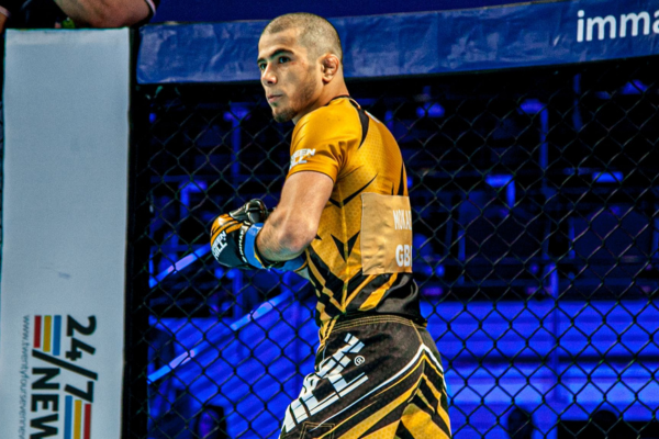 Alt-tekst: MMA-vechter in geel-zwarte rashguard en short staat gefocust in een kooi, klaar voor actie tijdens een professioneel gevecht, met een blauw verlichte arena op de achtergrond.