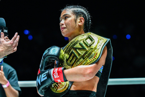 Een vrouwelijke kickbokser staat in de ring met gevlochten haar en bokshandschoenen aan, terwijl ze trots een grote gouden kampioensgordel van ONE om haar schouders draagt.