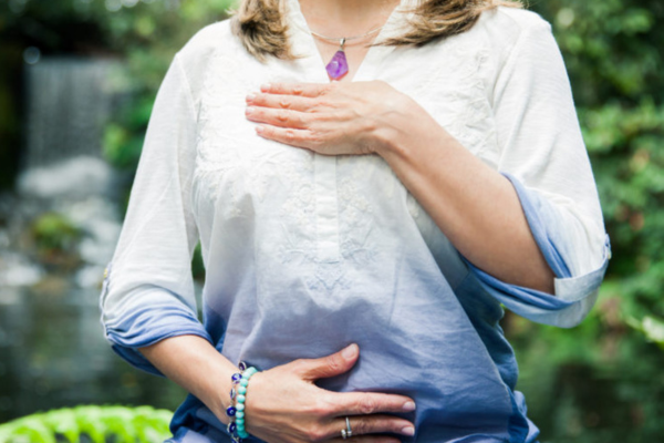 Vrouw die in meditatiehouding zit met één hand op haar hart en de andere op haar buik, in een natuurlijke buitenomgeving.