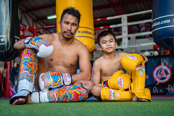 Muay Thai-vechter oefent een krachtige knietrap op een trainer met pads in een open trainingsruimte, omringd door natuur en zonlicht.