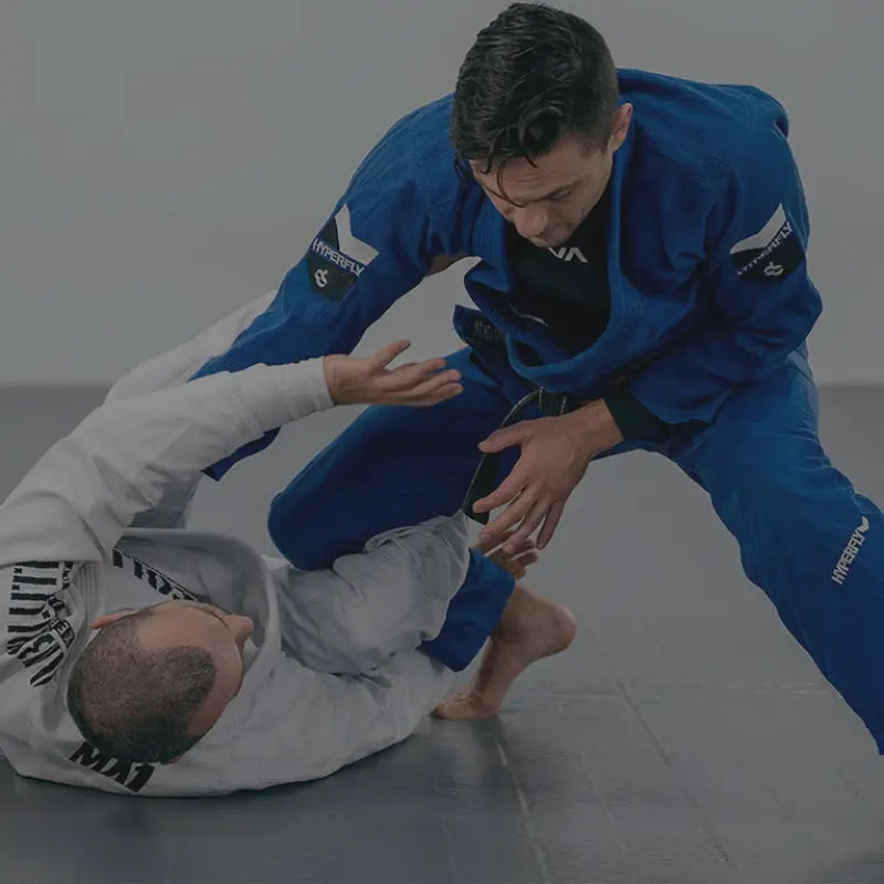 Twee mannen trainen Braziliaans Jiu Jitsu op een mat; de ene draagt een blauwe gi en staat, terwijl de andere in een witte gi op de grond ligt en probeert te verdedigen.