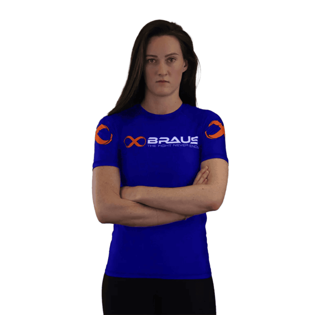 Een afbeelding van de voorkant van een blauwe damesrashguard met korte mouwen van Braus. Het shirt heeft oranje logo's en de tekst "Braus - The Fight Never Ends". Een vrouw staat met gekruiste armen en kijkt recht naar de camera.