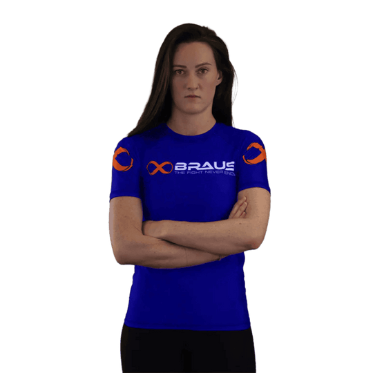 Een afbeelding van de voorkant van een blauwe damesrashguard met korte mouwen van Braus. Het shirt heeft oranje logo's en de tekst "Braus - The Fight Never Ends". Een vrouw staat met gekruiste armen en kijkt recht naar de camera.