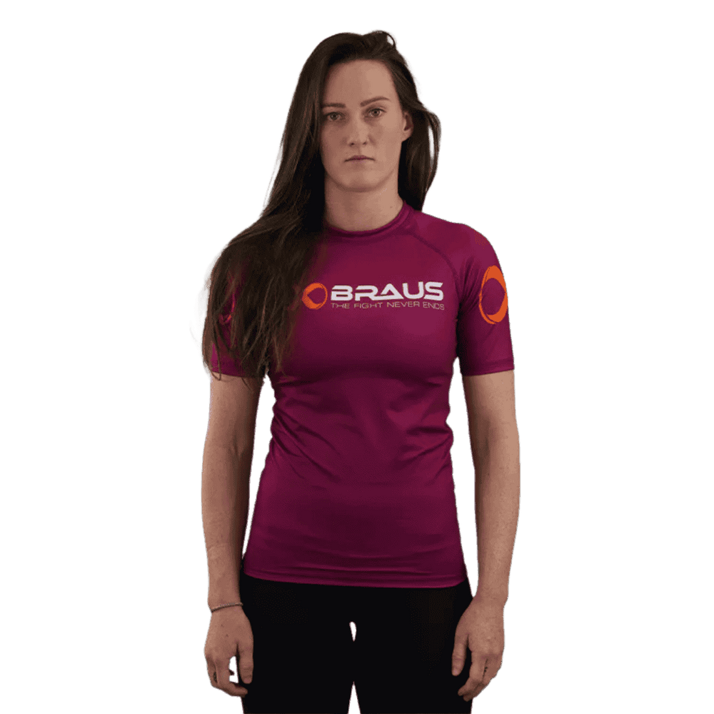 Een afbeelding van de voorkant van een paars damesrashguard met korte mouwen van Braus. Het shirt heeft een oranje logo en de tekst "Braus - The Fight Never Ends." De vrouw staat recht en kijkt direct naar de camera.