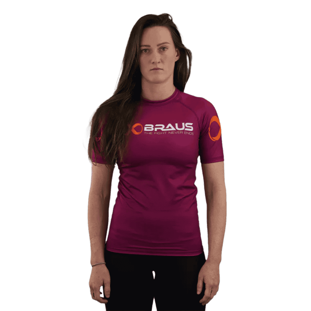 Een afbeelding van de voorkant van een paars damesrashguard met korte mouwen van Braus. Het shirt heeft een oranje logo en de tekst "Braus - The Fight Never Ends." De vrouw staat recht en kijkt direct naar de camera.