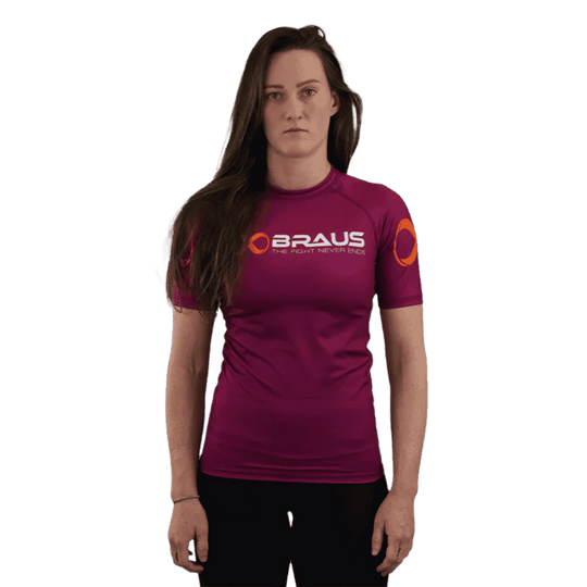 Een afbeelding van de voorkant van een paars damesrashguard met korte mouwen van Braus. Het shirt heeft een oranje logo en de tekst "Braus - The Fight Never Ends." De vrouw staat recht en kijkt direct naar de camera.