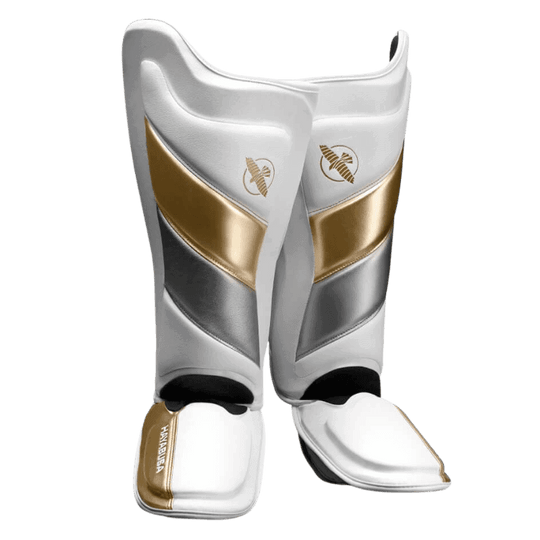 Een paar Hayabusa T3 Striking Scheenbeschermers in wit en goud, van voren bekeken. Ze hebben een strak ontwerp met het gouden Hayabusa valk-logo bovenop.