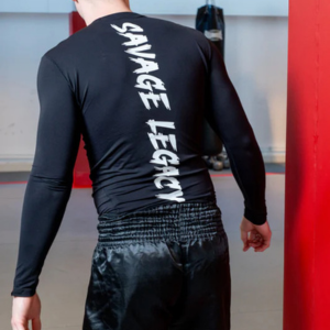 Man draagt een zwarte rash guard met lange mouwen met witte tekst "Savage Legacy" op de rug, staat in een gym met rode bokszakken op de achtergrond