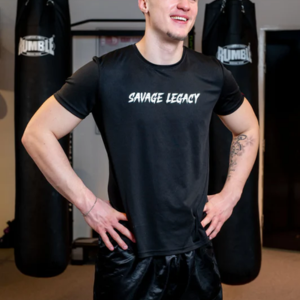 Man draagt een zwart T-shirt met witte opdruk "Savage Legacy" op de borst, staat ontspannen met handen op heupen, bokszakken zichtbaar op achtergrond