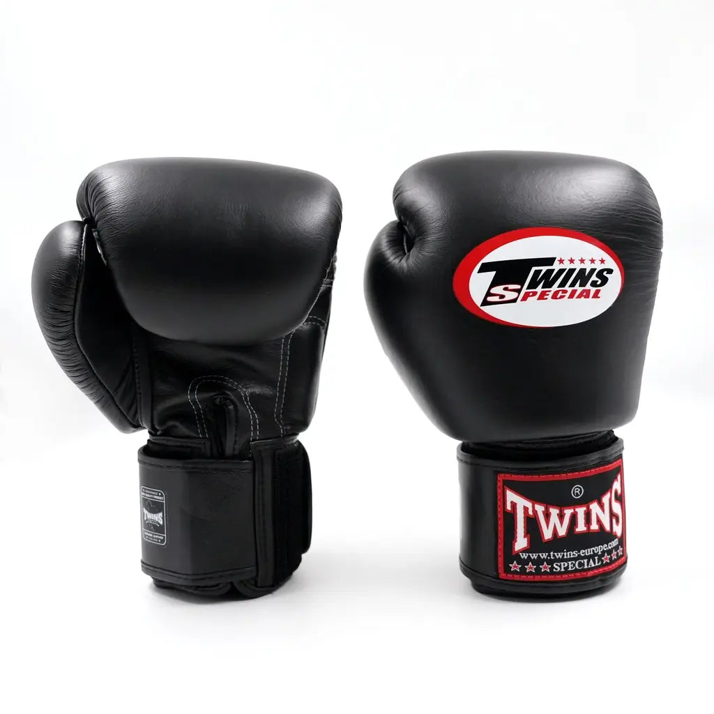 Zwarte Twins Special bokshandschoenen op witte achtergrond, één met zichtbare handpalmzijde en klittenbandsluiting, de ander met rode Twins-logo op de vuistzijde.