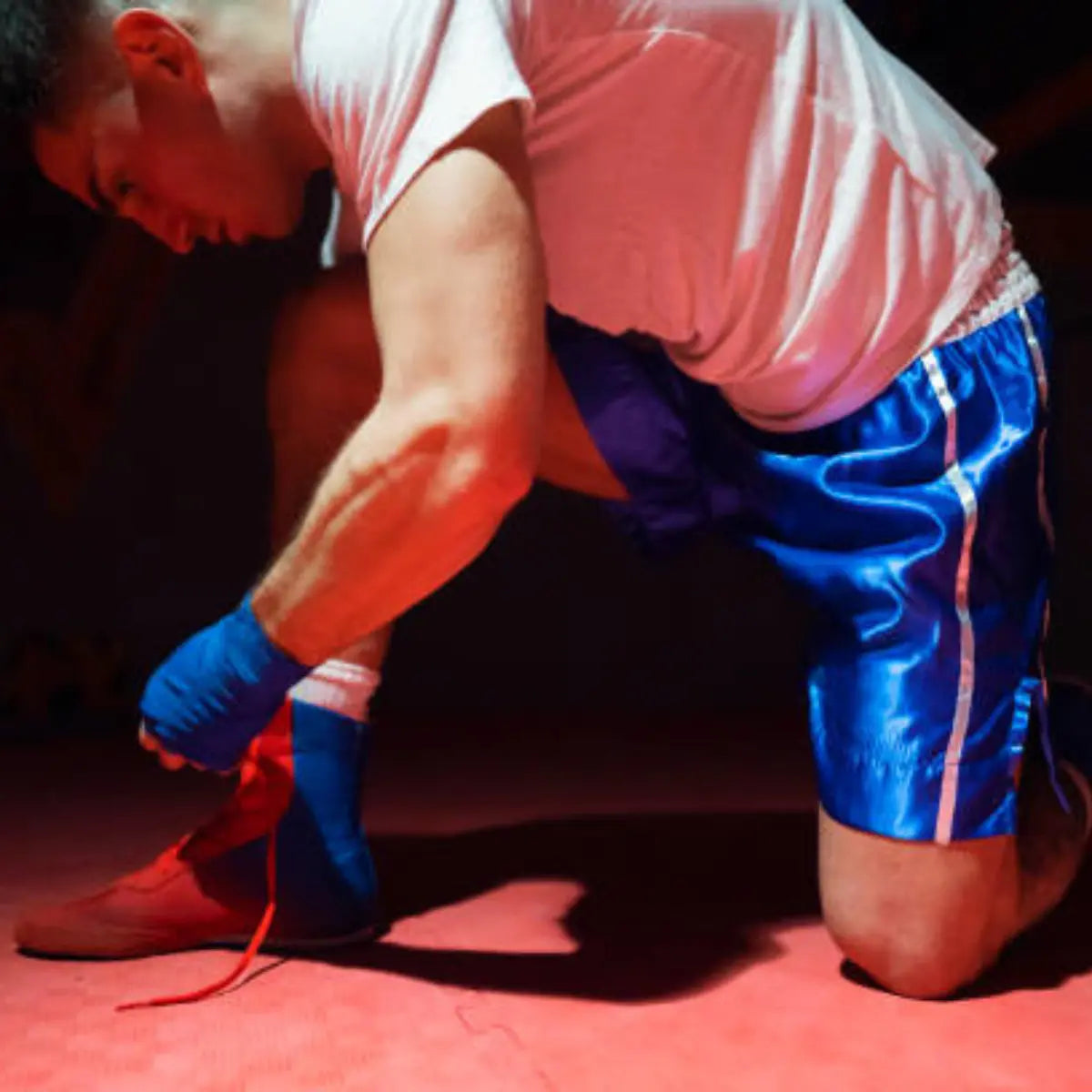 Bokser knielt op de mat terwijl hij zijn boksschoen strikt, gekleed in blauwe shorts en met bandages om zijn handen.