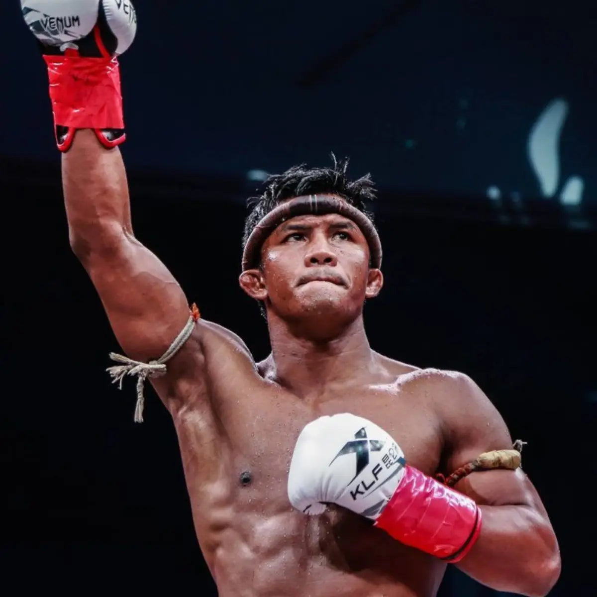 Muay Thai-legende Buakaw poseert krachtig in de ring met één vuist in de lucht, gekleed in zwarte shorts met zijn naam en traditionele armbanden.