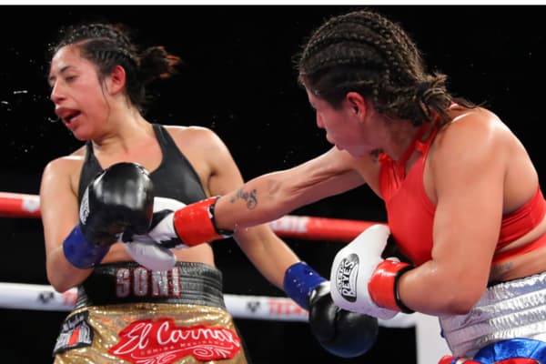 Twee vrouwelijke boksers in actie tijdens een wedstrijd, waarbij één vechter een krachtige stoot uitdeelt aan haar tegenstander. De scène toont focus, techniek en intensiteit in het vrouwenboksen.