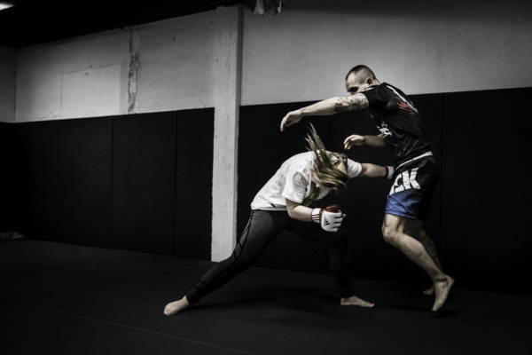 Twee MMA-beoefenaars trainen takedowns op een zwarte mat; een vrouw in witte shirt duikt laag in op haar tegenstander terwijl hij probeert balans te houden tijdens de oefening.
