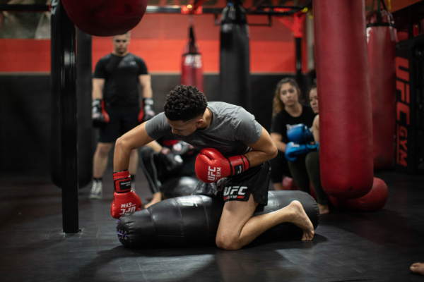 Man met rode UFC-handschoenen oefent ground-and-pound technieken op een grappling dummy in een UFC Gym, terwijl andere sporters toekijken en trainen in de achtergrond.