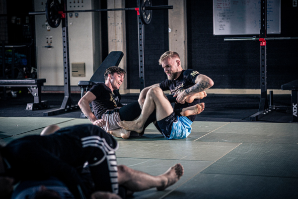 Twee mannelijke grapplers oefenen beenklemtechnieken op de mat tijdens een Brazilian Jiu-Jitsu training, lachend en ontspannen in een sportschool met krachttrainingsapparatuur op de achtergrond.
