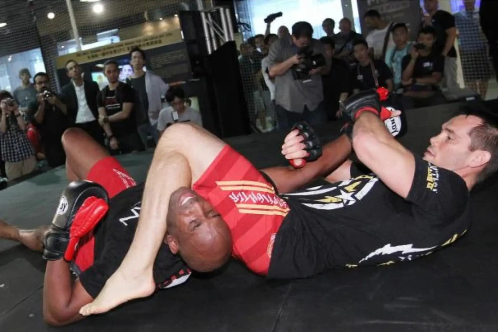 Twee vechters op de grond tijdens een grapplingdemonstratie, waarbij één vechter een armbar inzet terwijl toeschouwers rondom kijken.