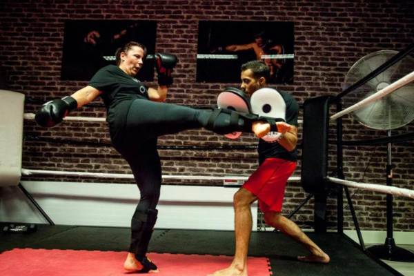 Vrouw in zwarte sportkleding voert een hoge trap uit tijdens een kickbokstraining, terwijl een trainer met focus pads de trap opvangt in een sportschool met een ring en bakstenen muur.