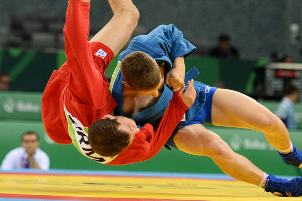 Twee mannelijke sambo-vechters in actie op de mat; de vechter in blauw werpt zijn tegenstander in rood met een dynamische worp tijdens een officiële wedstrijd.