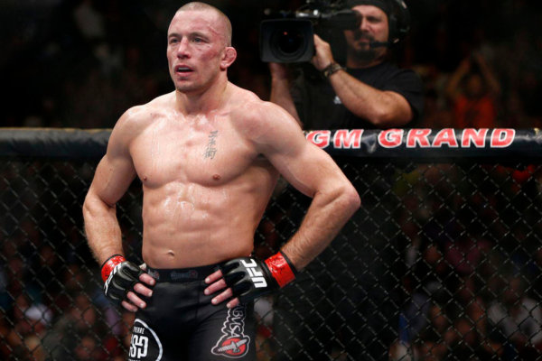 Gespierde MMA-vechter staat bezweet in de kooi met handen op zijn heupen na een intens gevecht, terwijl een cameraman op de achtergrond filmt in de MGM Grand Arena.