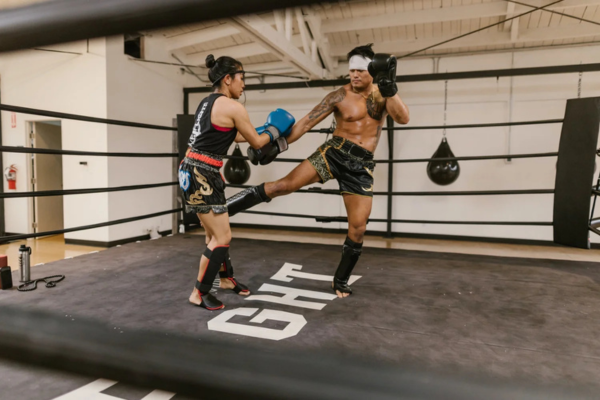 Twee Muay Thai-vechters trainen in een boksschool; de man voert een low kick uit richting de vrouw, beiden dragen scheenbeschermers, handschoenen en traditionele shorts in een sparringring.