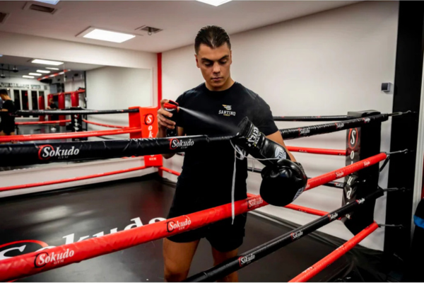 Alt tekst: Man in zwarte sportoutfit maakt zijn bokshandschoenen vast aan de touwen van een rood-zwarte boksring in een moderne gym.