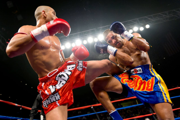 Twee Muay Thai-vechters in actie waarbij de ene een trap uitdeelt en de ander zich verdedigt in een fel verlichte boksring.