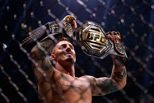 UFC-vechter houdt trots een kampioensgordel boven zijn hoofd terwijl hij in de kooi staat, met zichtbare tatoeages en gefocuste blik onder het licht van de arena.