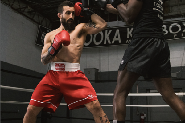 Twee boksers trainen in een sportschool; de vechter in rode short verdedigt zich tegen een aanval van zijn sparringpartner.