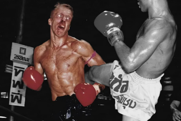 Muay Thai-vechter ontvangt een harde trap in het gezicht tijdens een gevecht; de impact is zichtbaar op zijn bebloede gezicht terwijl zijn tegenstander krachtig uithaalt met rode bokshandschoenen en traditionele Muay Thai-short.