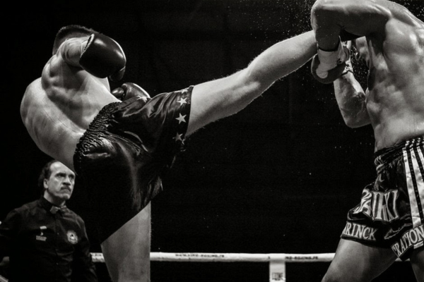 Zwart-witfoto van een kickbokswedstrijd waarbij een vechter een hoge trap uitdeelt, met een scheidsrechter die toekijkt in de ring.