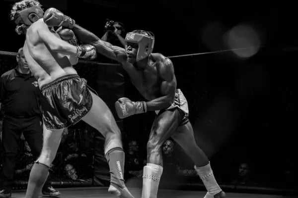 Twee kickboksers in de ring tijdens een intens gevecht, waarbij één vechter een krachtige stoot uitdeelt terwijl de ander achteruit wijkt, zwart-wit actiefoto.