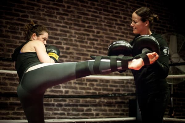 Vrouwelijke kickbokser voert een hoge trap uit tijdens training en draagt een zwarte scheenbeschermer voor bescherming van scheen en wreef. De focus ligt op veiligheid, techniek en gecontroleerde kracht in de gym.