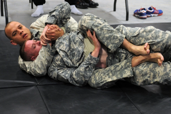 Twee militairen in camouflage-uniform oefenen een grondtechniek tijdens een vechtsporttraining op een mat.