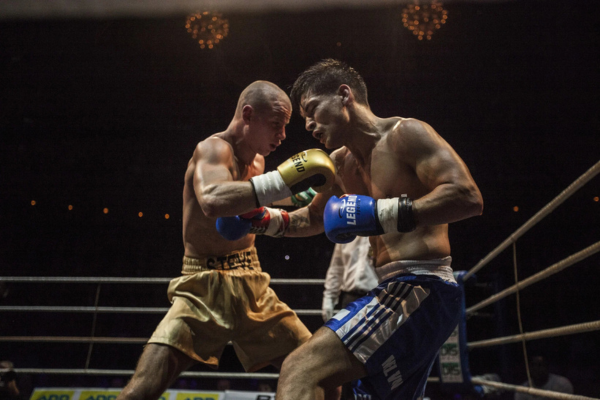 Twee boksers in de ring wisselen krachtige stoten uit tijdens een professioneel gevecht, met een scheidsrechter op de achtergrond en fel licht dat de actie benadrukt.