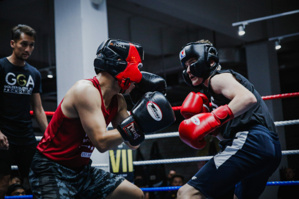 Twee jonge boksers met hoofdbescherming en rode bokshandschoenen sparren intensief in een bokswedstrijd, terwijl een trainer of scheidsrechter toekijkt in de ring.