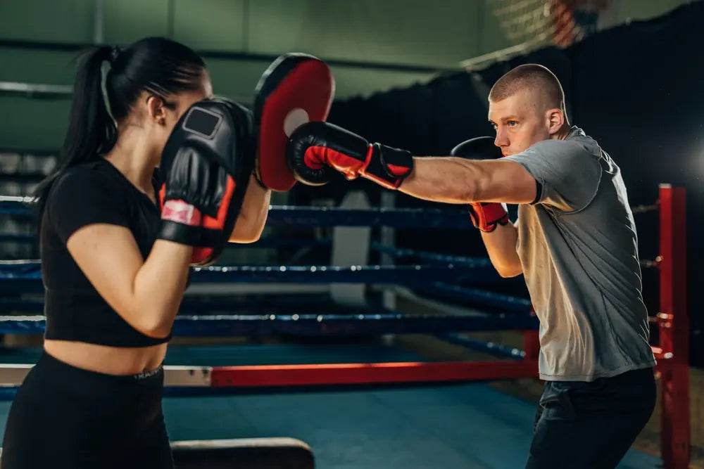 Een man en een vrouw trainen samen in een boksschool. De man draagt bokshandschoenen en oefent stoottechniek op focus mitts die de vrouw vasthoudt. Ze staan tegenover elkaar in een boksring, gefocust en in actie tijdens een vechtsporttraining.