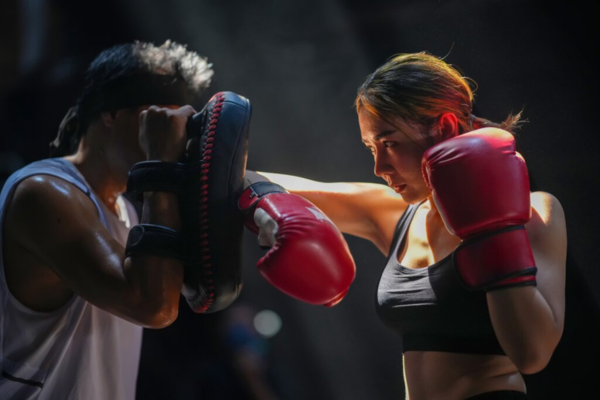 Vrouw met rode bokshandschoenen traint gericht op focus pads tijdens een intensieve bokssessie, met een trainer die de pads vasthoudt in een donkere sportschoolomgeving.