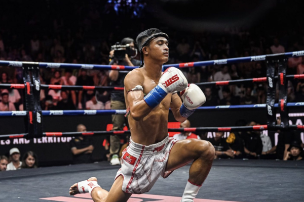 Muay Thai-vechter knielt in het midden van de ring tijdens het uitvoeren van de Wai Kru Ram Muay-ceremonie, met publiek op de achtergrond.