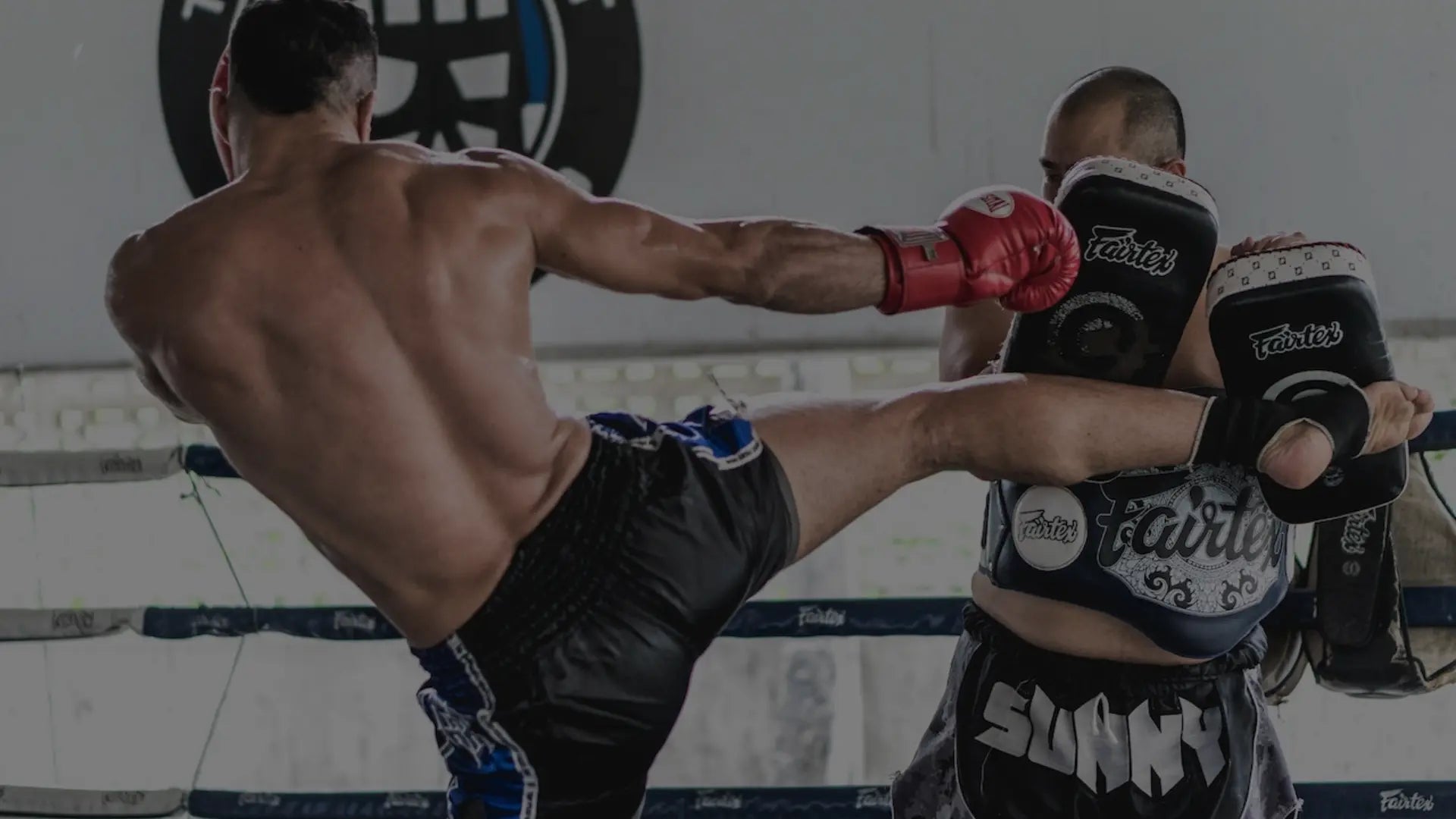 Een vechter geeft een hoge trap terwijl de trainer Fairtex trapkussens en bodyprotectie vasthoudt tijdens een intensieve Muay Thai sessie.
