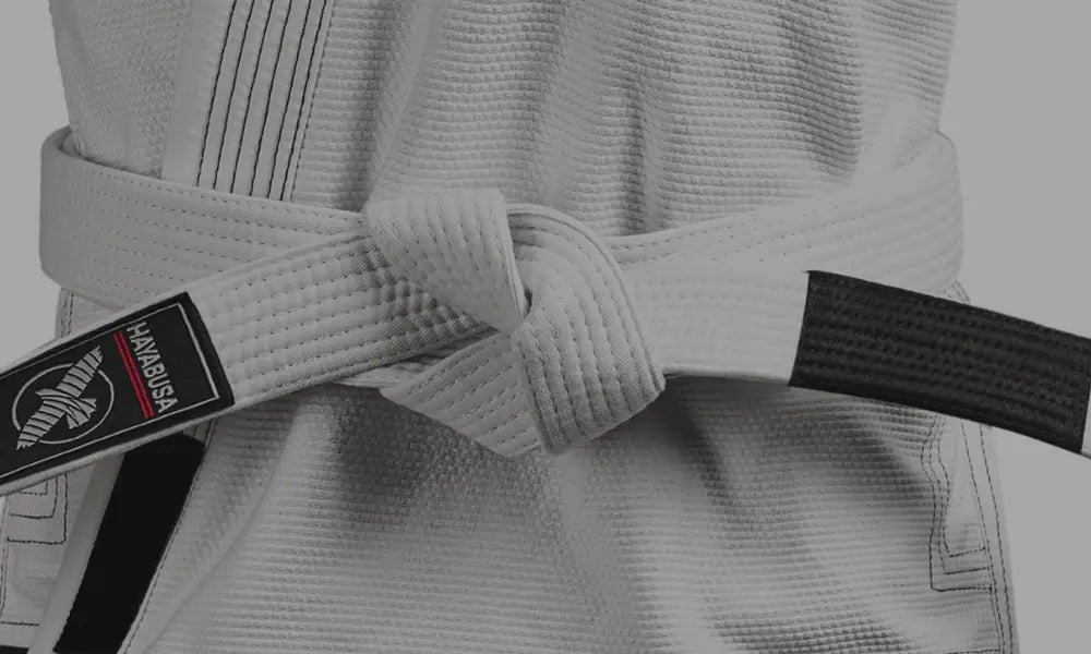 Close-up van een witte BJJ-band om een gi, met zichtbaar Hayabusa-label en zwarte eindstrook, strak gestrikt rond het middel.