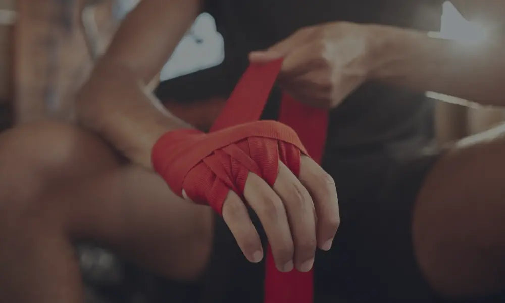Close-up van een hand die wordt ingepakt met rode bokshandschoenen bandages door een trainer, in een gymomgeving.
