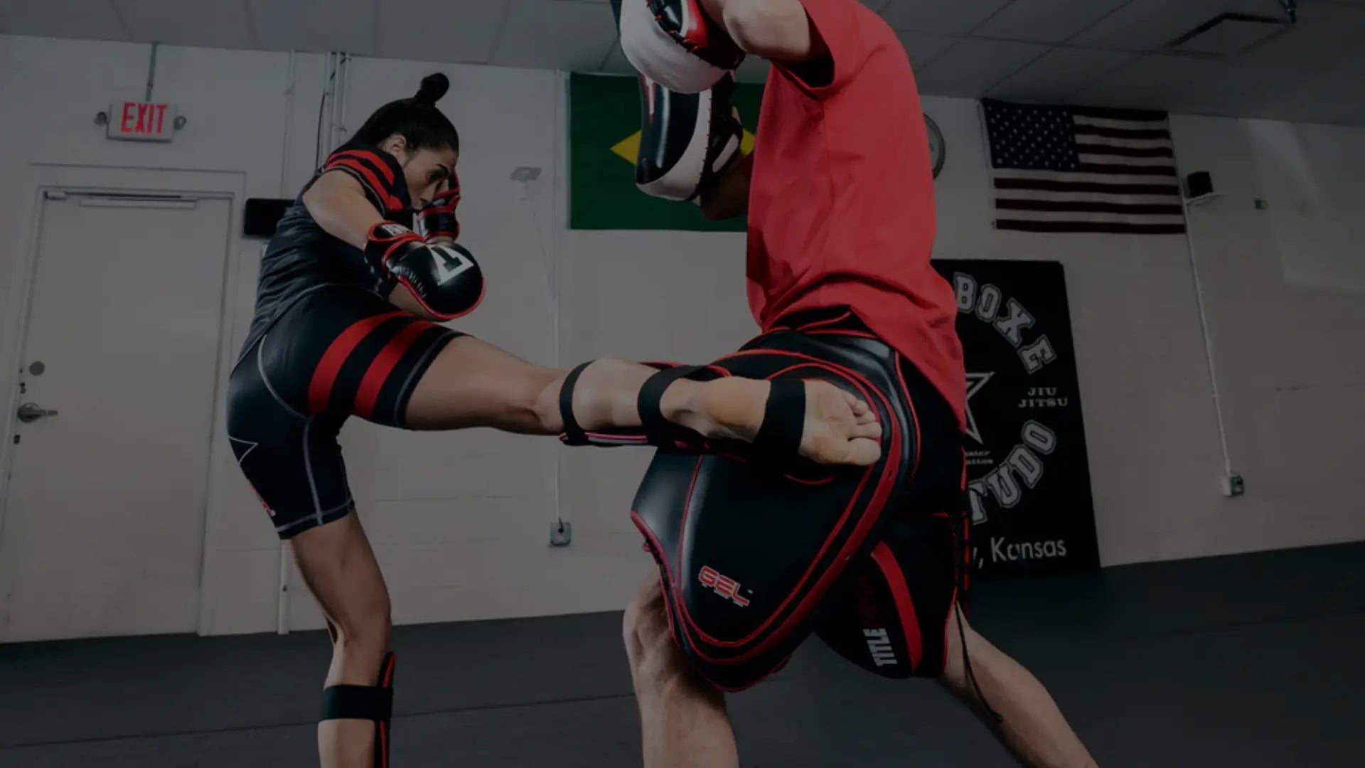 Een sporter geeft een harde trap terwijl ze beenbeschermers draagt; de trainer vangt de impact op met dikke zwarte en rode trapkussens in een trainingsruimte.