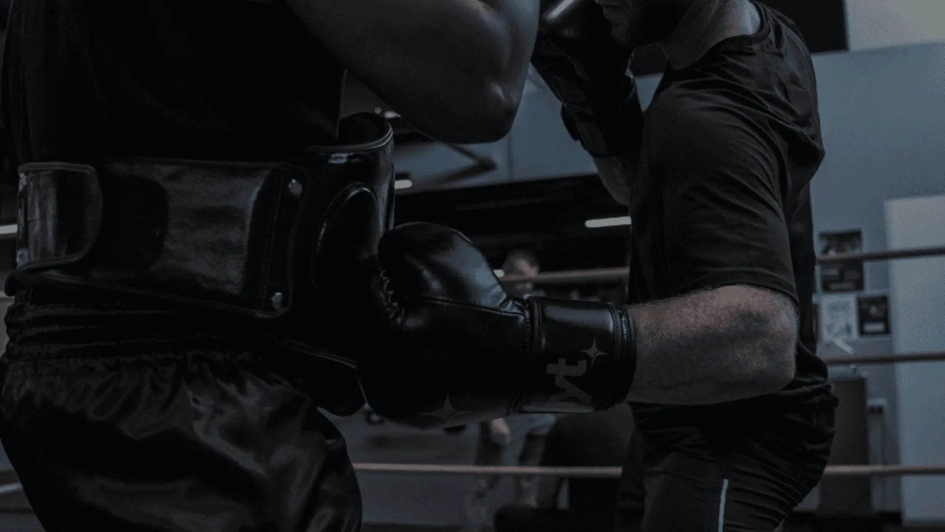 Twee sporters oefenen bokscombinaties waarbij één persoon een bodyprotector draagt en de ander met zwarte bokshandschoenen bodyshots traint.