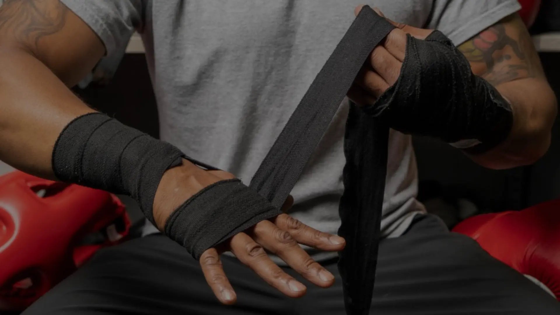 Close-up van een sporter die zwarte boksbandages strak om zijn handen wikkelt ter voorbereiding op een bokstraining, met rode beschermers op de achtergrond.