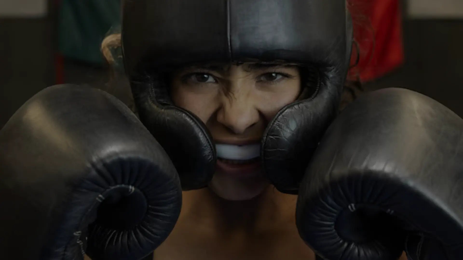 Close-up van een bokser met zwarte handschoenen en hoofdbeschermer die een wit bitje toont tijdens een intense trainingsmoment.